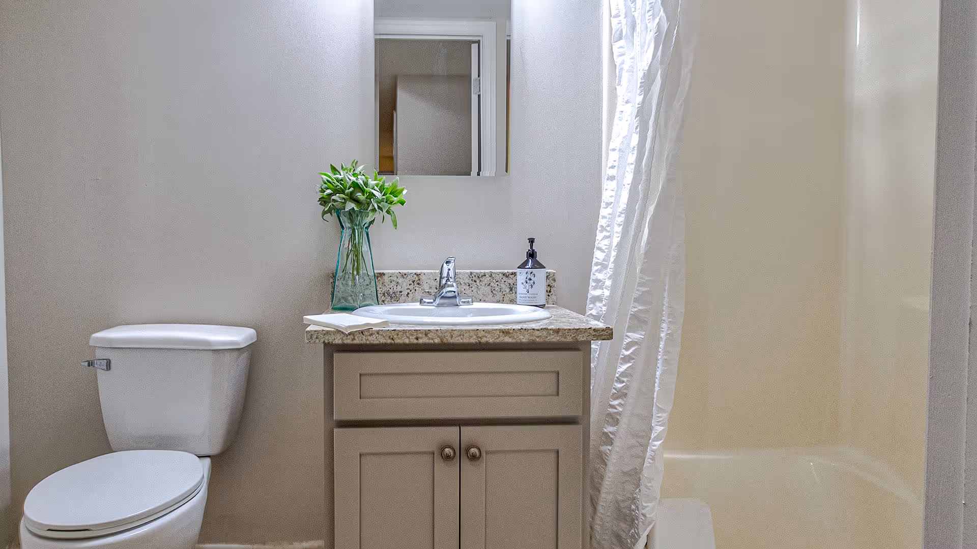 A clean bathroom featuring a white toilet, a vanity with a granite countertop and a sink, a small mirror above the sink, a green vase with leafy plants, a soap dispenser, and a white shower curtain partially covering a bathtub.