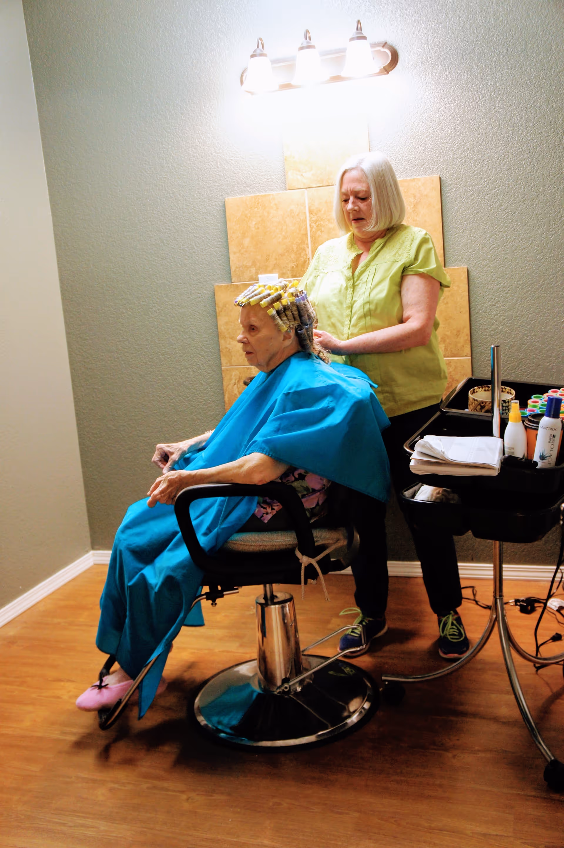 An elderly woman with hair rollers sits in a salon chair covered with a blue cape while a hairstylist stands behind her, working on her hair in a room with wooden flooring and a wall-mounted light fixture.