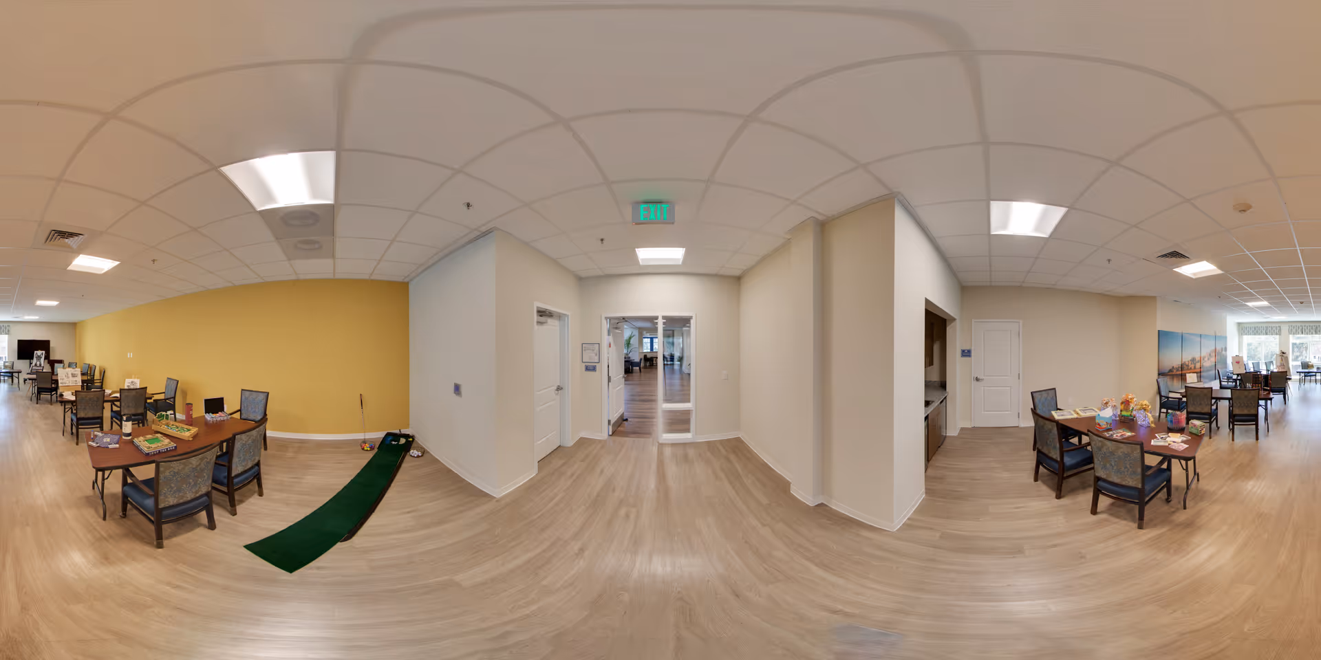 Wide panoramic view of a spacious, well-lit common area in an assisted living facility. The room features light wood flooring, beige and yellow walls, and several tables with chairs arranged for activities or dining. There is a small indoor putting green on the floor near one table. The ceiling has recessed lighting and a visible exit sign above a doorway leading to another room.