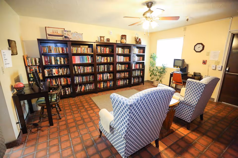 A cozy reading room with three large dark wood bookshelves filled with books against the wall. Two patterned armchairs face a small wooden table in the center of the room. There is a ceiling fan with lights above, a window letting in natural light, a potted plant near the window, and a small desk with a computer and chair in the corner. The floor is covered with reddish-brown tiles.