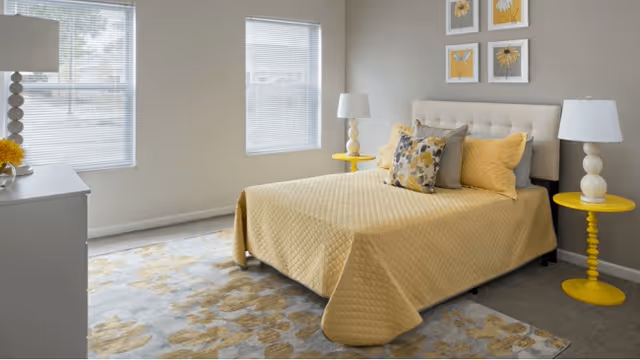 A bedroom with a bed covered in a yellow quilt and multiple pillows, two yellow round side tables each with a white lamp, a white dresser with a lamp and flowers, two windows with blinds, and four framed floral artworks on the wall above the bed.