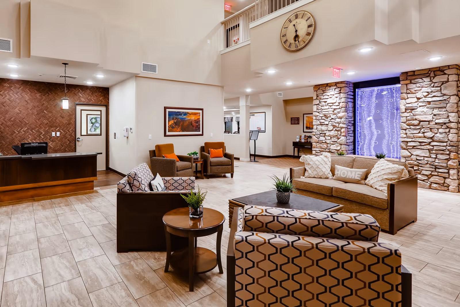 A spacious and well-lit living room area in a senior living facility featuring a reception desk on the left, multiple comfortable chairs and sofas with patterned and solid cushions, a large wall clock, stone accent walls, and a decorative water feature illuminated with blue lighting.