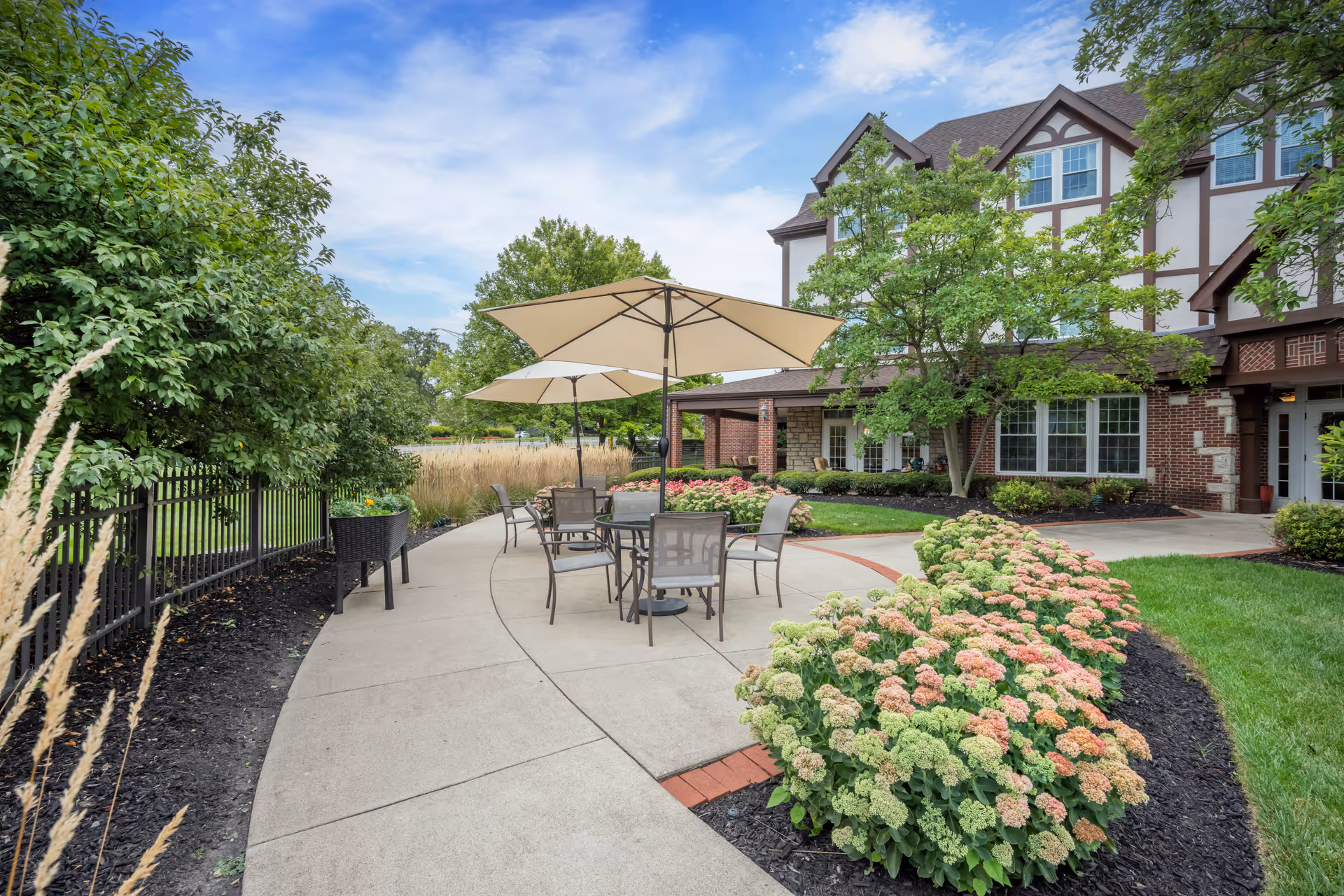 Outdoor patio with tables, umbrellas, and flowerbeds in front of a multi-story senior living building.