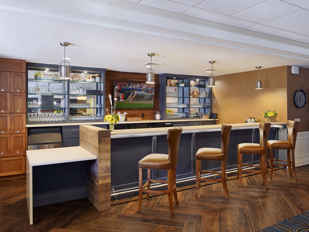 Interior view of a modern bar area with four brown leather bar stools lined up along a white countertop. Behind the bar are shelves with glassware and decorative items, a mounted flat-screen TV showing a baseball game, and wooden cabinets. The floor has a herringbone wood pattern, and the walls are painted in warm tones with pendant lights hanging from the ceiling.
