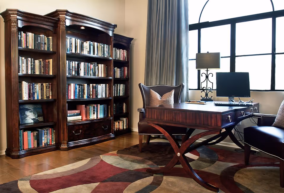 A cozy office or study room with a large wooden desk, two upholstered chairs, a computer monitor, a decorative lamp, and a tall wooden bookshelf filled with books. The room has a large window with a curtain and a patterned area rug on the wooden floor.
