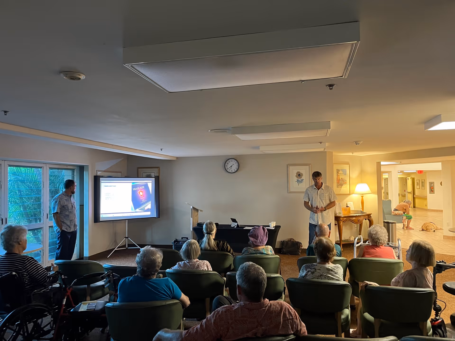 A group of elderly people seated in a common area watching a presentation. Two men stand at the front near a projector screen displaying a slide about fraud risk. The room has green chairs, a clock on the wall, framed pictures, and a lamp on a side table. In the background, a hallway is visible with a person sitting and a dog lying on the floor.