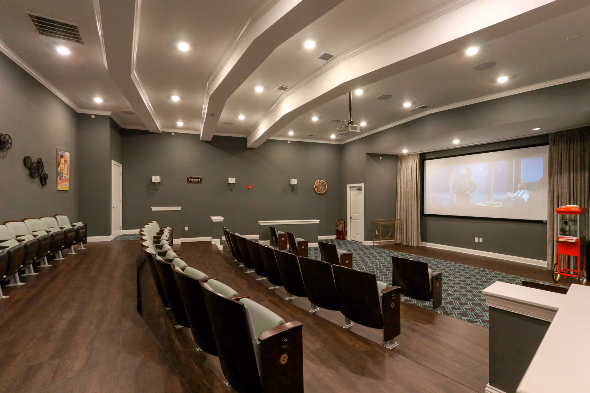 Interior community movie theater with rows of upholstered seats facing a large screen, recessed ceiling lights, and a popcorn machine.