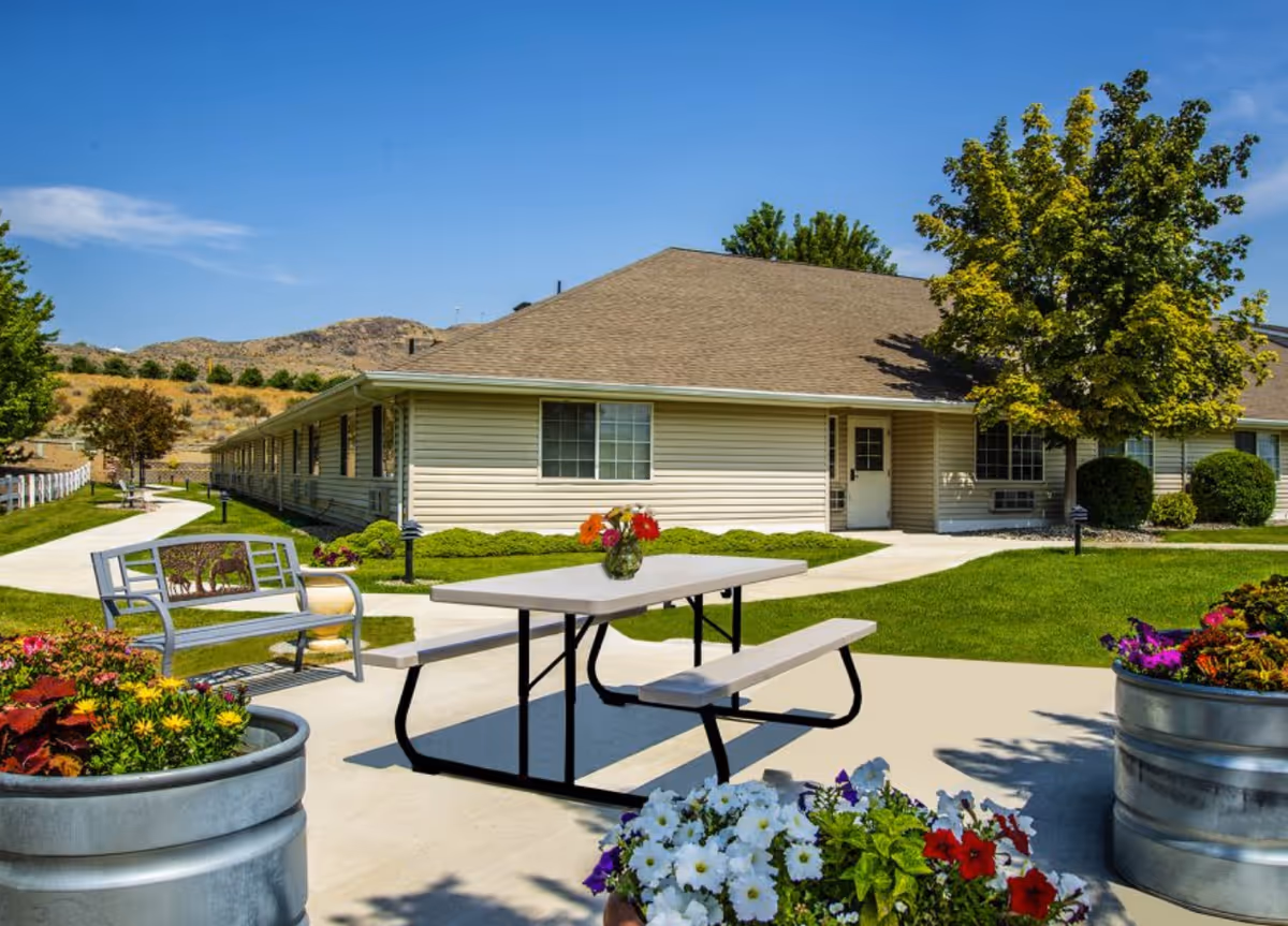 Outdoor patio area at Apple Springs Senior Living featuring a picnic table with a vase of flowers, a metal bench, large planters with colorful flowers, a well-maintained lawn, trees, and a single-story building under a clear blue sky.