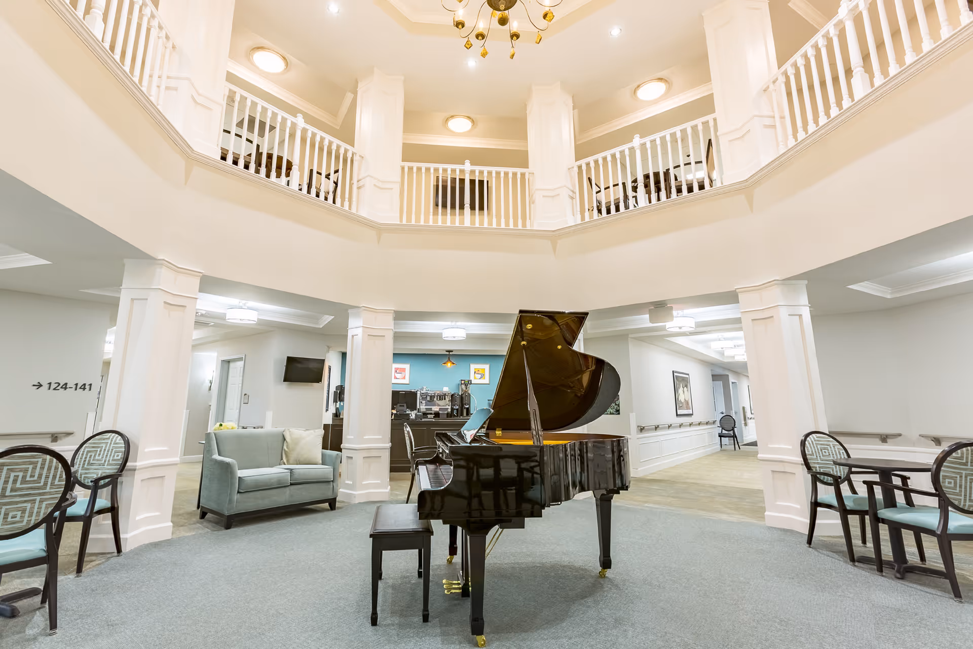 A spacious and elegant common area in a senior living facility featuring a black grand piano with an open lid in the center. Surrounding the piano are several seating arrangements including a light blue loveseat and multiple chairs with patterned cushions. The area is well-lit with ceiling lights and has white pillars and railings on the upper level, creating an open and airy atmosphere.
