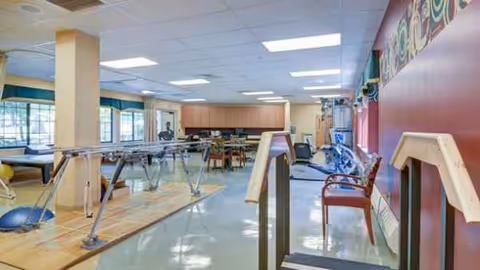 Interior senior living gym area with parallel bars, exercise equipment, and seating along a red wall.