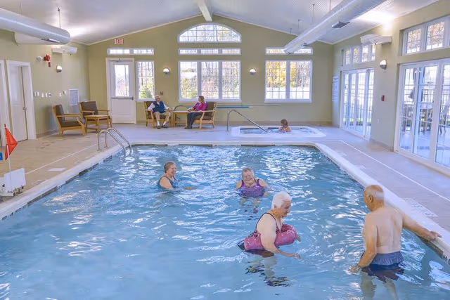 Indoor swimming pool area with four elderly people in the water and one person in a hot tub. Two elderly people are seated on chairs near large windows in the background. The room has high ceilings, large windows, and glass doors letting in natural light.