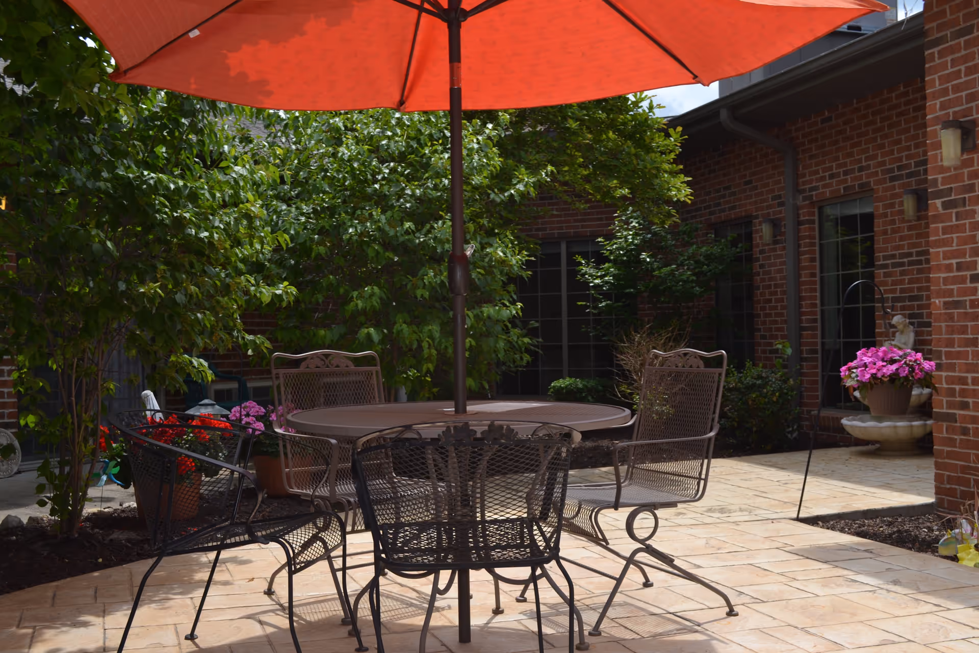 Outdoor patio area with a round metal table and four metal chairs under a large red umbrella. The patio is surrounded by greenery, potted flowers, and brick building walls with windows.