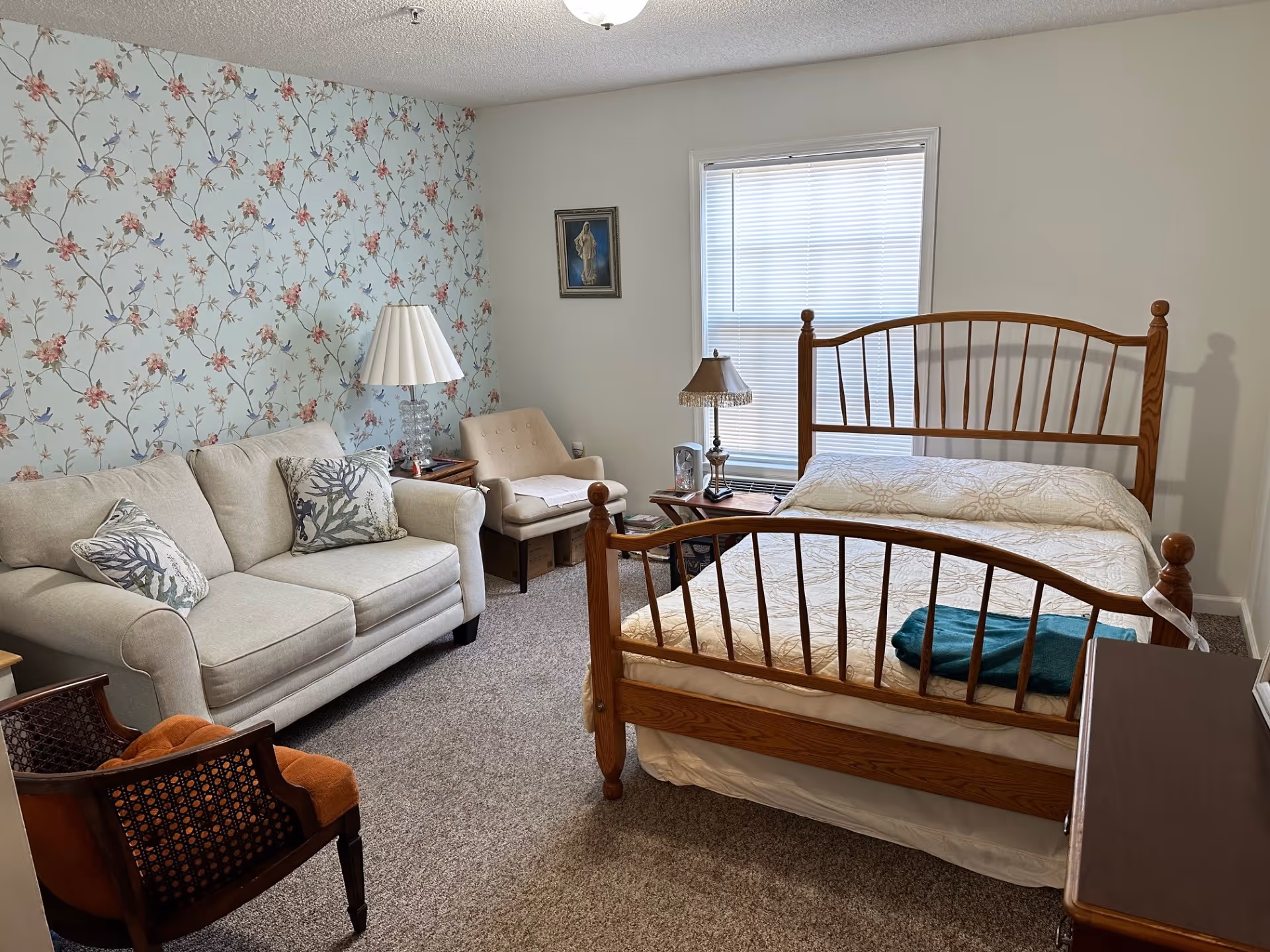 A cozy bedroom with a wooden bed frame and a beige bedspread. There is a beige loveseat with two decorative pillows, a beige armchair, and a wooden chair with an orange cushion. A side table with a lamp and a small table with another lamp are next to the bed. The wall behind the loveseat has floral wallpaper with pink flowers and blue birds, while the other walls are plain white. A window with closed blinds is above the bedside table, and a framed picture hangs on the wall.
