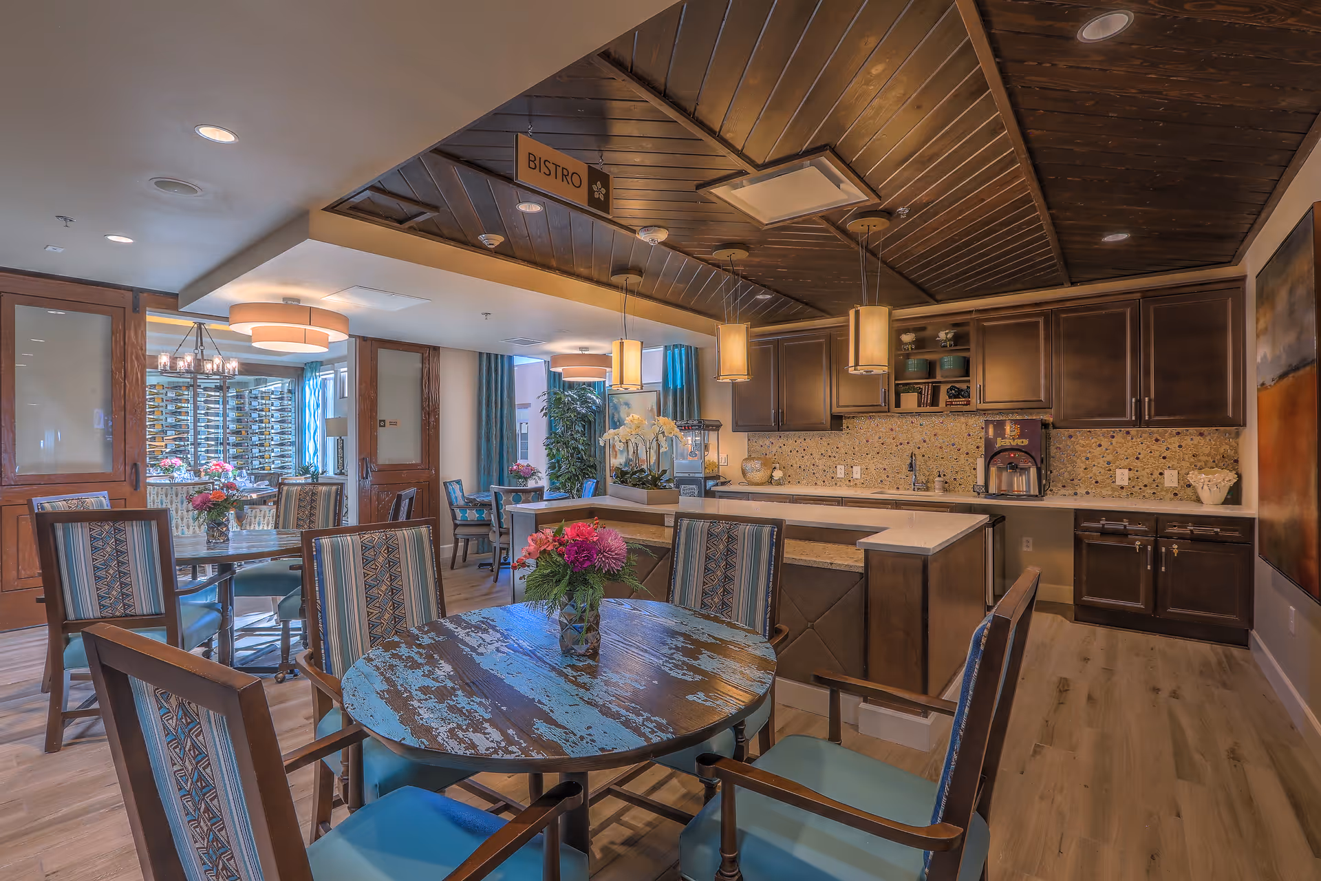 Interior view of a bistro area in MorningStar Assisted Living & Memory Care of Santa Fe featuring a round wooden table with a vase of flowers, surrounded by chairs with patterned upholstery. The background shows a kitchen area with dark wooden cabinets, a mosaic tile backsplash, pendant lights, and additional seating with tables and chairs near large windows with blue curtains.