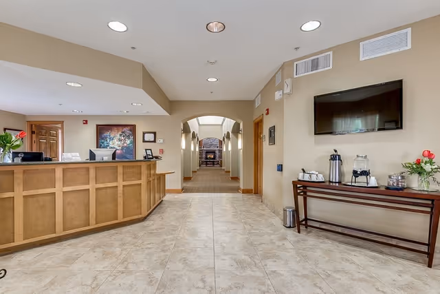 Reception area with a wooden front desk on the left and a hallway leading to a room with a fireplace in the background. On the right, there is a table with a water dispenser, coffee thermos, cups, and flowers under a mounted flat-screen TV. The floor is tiled, and the walls are painted beige.