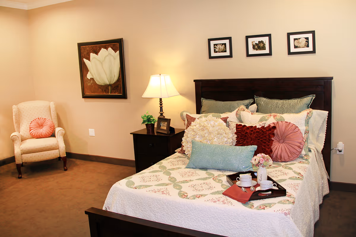 A cozy bedroom in a senior living facility featuring a bed with a floral quilt and multiple decorative pillows, a wooden nightstand with a lamp, a small plant, and a framed photo. There is a comfortable armchair with a round pink cushion in the corner, and floral artwork hanging on the beige walls.