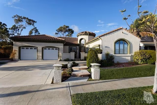 Exterior view of a single-story residential building with a tiled roof, two garage doors, a driveway, and a landscaped front yard with shrubs and trees under a clear blue sky.