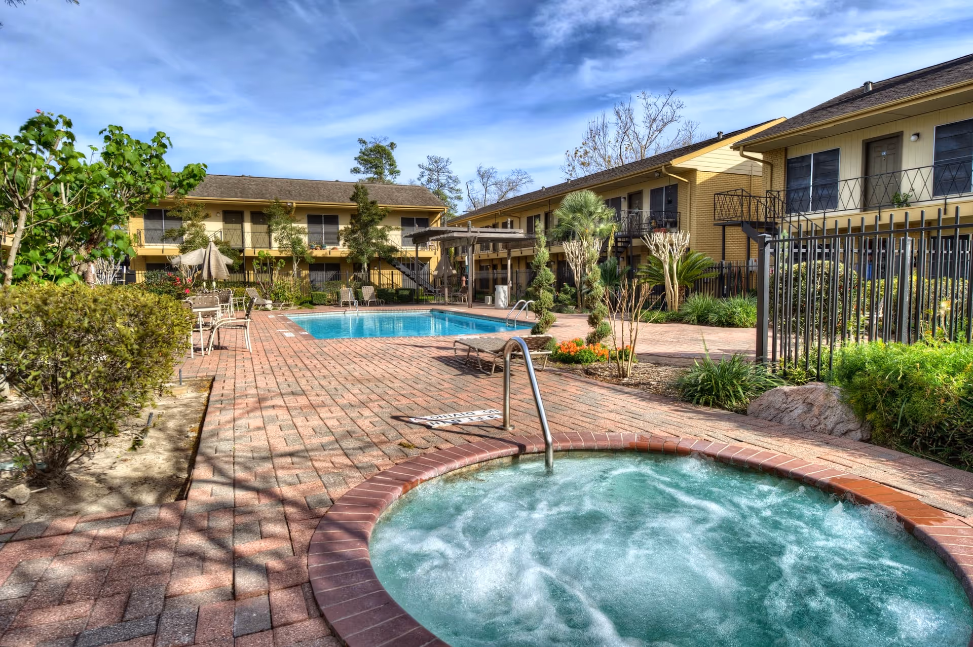Outdoor view of a retirement community featuring a bubbling hot tub in the foreground, a swimming pool surrounded by lounge chairs and tables with umbrellas, and two-story residential buildings with balconies in the background under a partly cloudy sky.