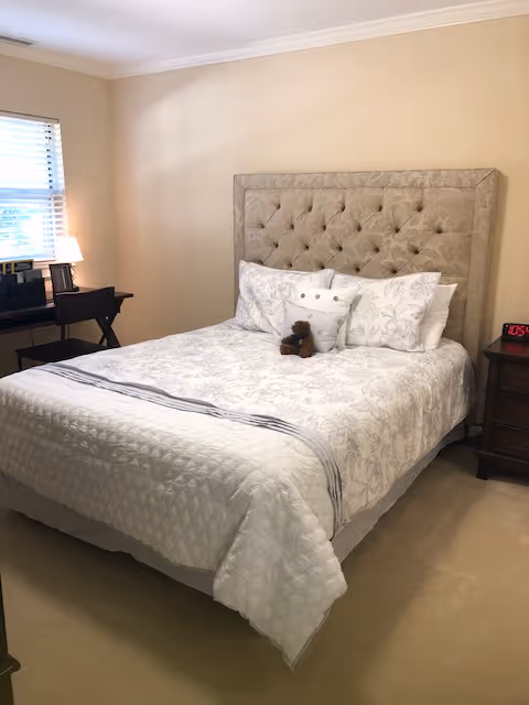 A neatly made bed with a tufted headboard, nightstand, lamp, window, and a small teddy bear on the pillows.