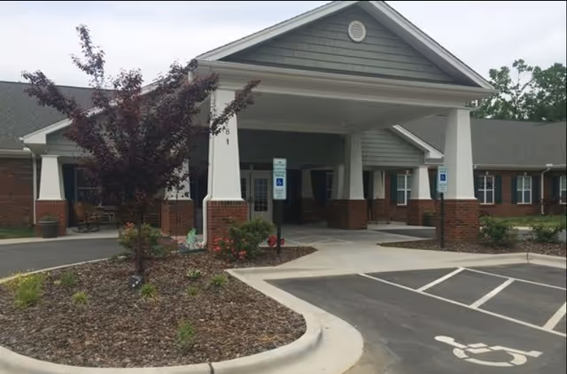 Entrance of a single-story assisted living facility with a covered drop-off area supported by white columns with brick bases. There are two handicap parking spaces in front, a small landscaped area with a tree and plants, and the building has green siding with a gray roof.