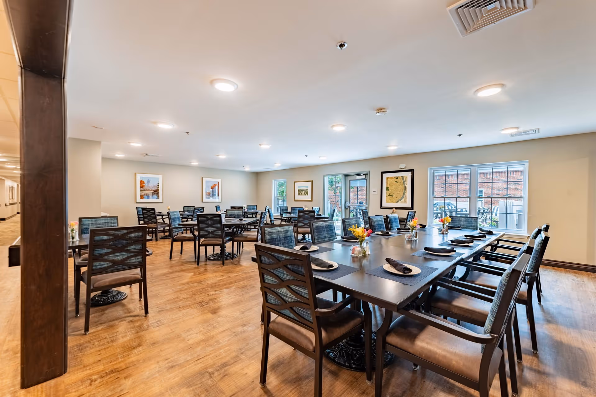 A spacious dining room in a senior living facility with multiple tables and chairs arranged neatly. The tables are set with placemats, napkins, and small flower arrangements. The room has wooden flooring, light-colored walls, framed artwork, and large windows letting in natural light. There is a door leading to an outdoor area visible in the background.