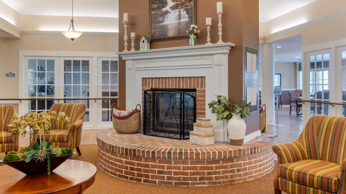 Cozy community lounge with a brick-hearth fireplace, striped armchairs, and a round table with a floral arrangement.