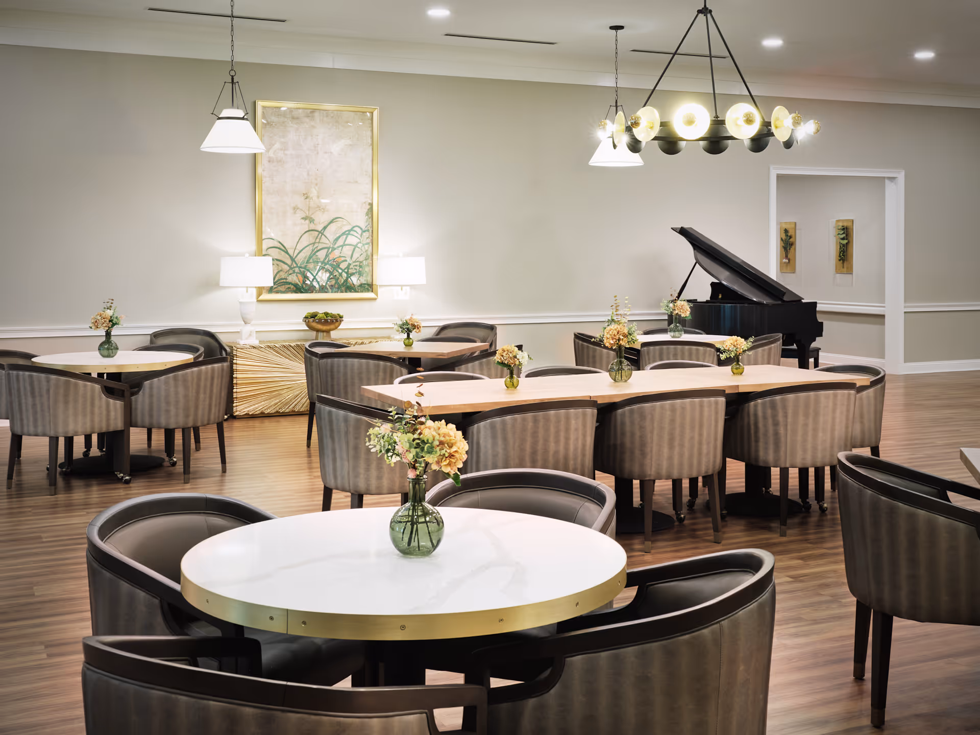 A senior living facility dining room with multiple tables and chairs arranged neatly. Each table has a small vase with flowers. The room features wooden flooring, a grand piano in the corner, wall art, and modern light fixtures hanging from the ceiling.