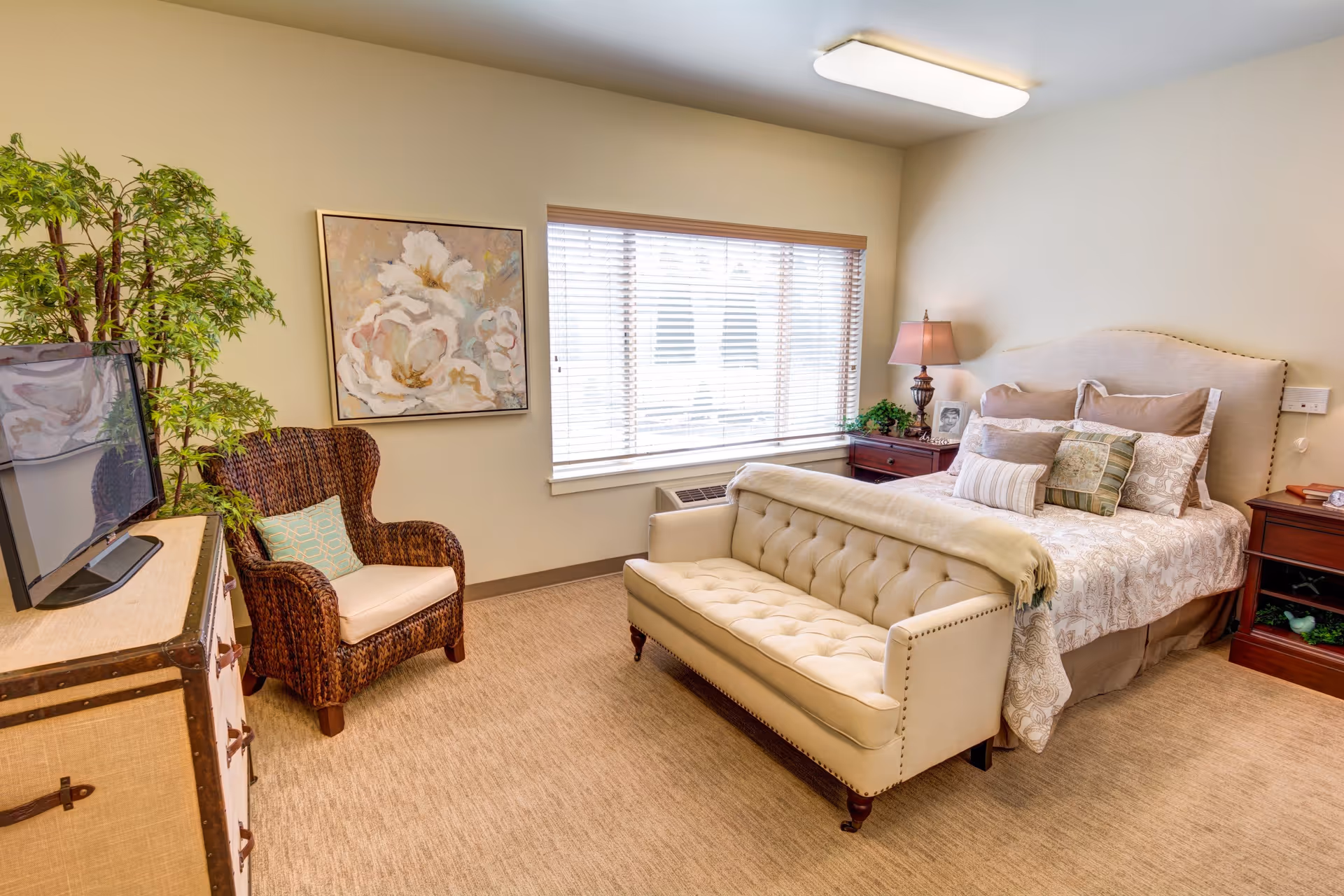 A cozy bedroom in an assisted living facility featuring a bed with multiple pillows and a beige tufted bench at the foot. There is a wooden nightstand with a lamp and framed photo, a large window with blinds, a wicker armchair with a cushion, a TV on a dresser, and a large floral painting on the wall. The room has beige carpeting and neutral-colored walls.