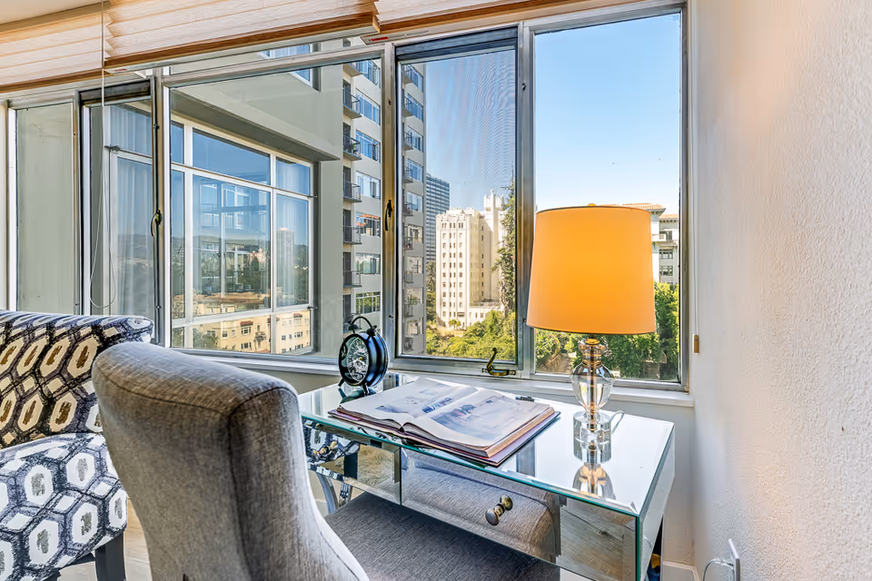 A bright interior corner with a glass-top desk holding an open book, a decorative clock, and a lit table lamp. Next to the desk is a patterned armchair and a gray upholstered chair. Large windows provide a view of nearby buildings and greenery outside.