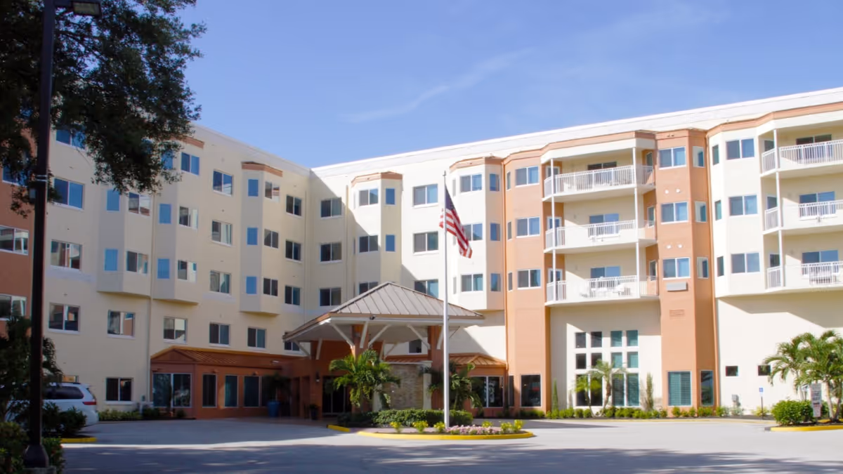 Exterior view of a multi-story senior living facility building with balconies, numerous windows, a covered entrance, and an American flag on a flagpole in front. There are palm trees and landscaping around the entrance area under a clear blue sky.