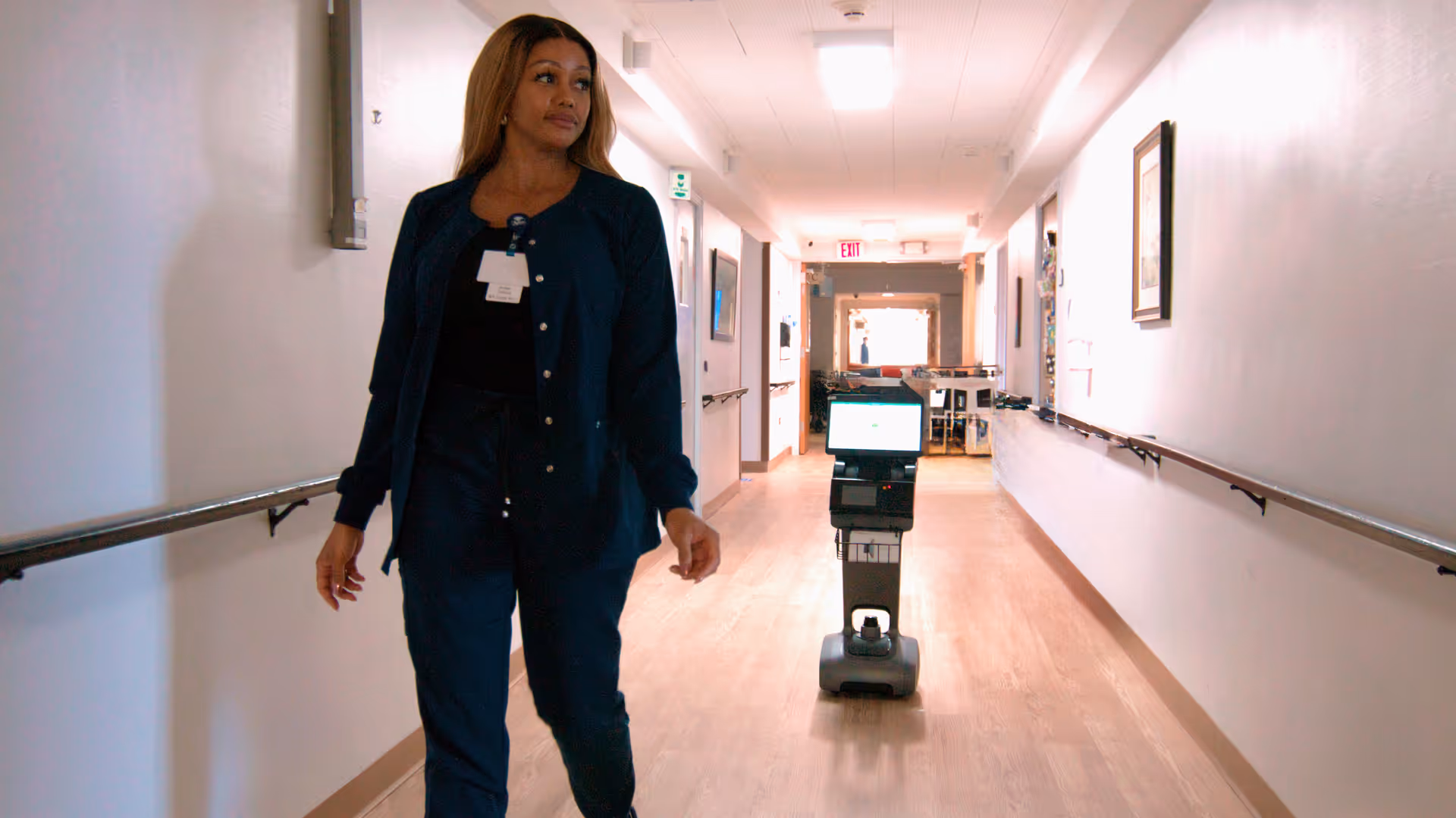 A woman in dark blue scrubs walking down a well-lit hallway with wooden floors and handrails on both sides. A small robot with a screen is positioned behind her in the hallway.