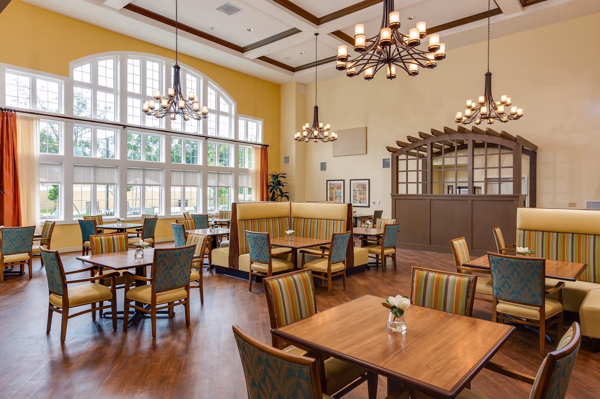 A spacious dining room with large arched windows letting in natural light. The room features wooden tables with a mix of striped and patterned upholstered chairs and booth seating. Elegant chandeliers hang from a coffered ceiling, and small flower vases decorate each table. The walls are painted yellow and beige, creating a warm and inviting atmosphere.