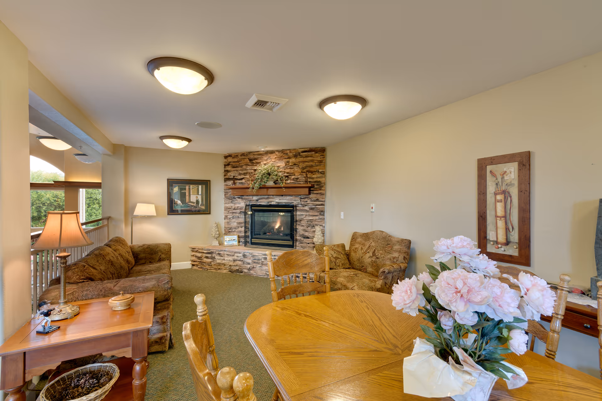 A cozy living room area with two patterned sofas facing a stone fireplace with a wooden mantel decorated with greenery. A wooden dining table with chairs and a vase of pink flowers is in the foreground. The room has beige walls, carpeted floor, and ceiling lights. A framed picture and a floor lamp are visible near one sofa.