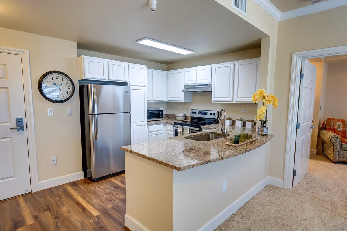 Modern kitchen with white cabinets, granite countertops, stainless steel refrigerator and stove, a sink on the curved countertop, and a clock on the wall. Adjacent room with a plaid armchair is visible through an open door.