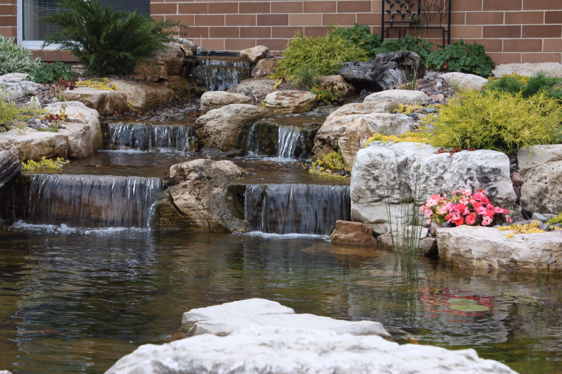 A landscaped garden pond with tiered rock waterfalls, surrounding plants and flowers in front of a brick building.