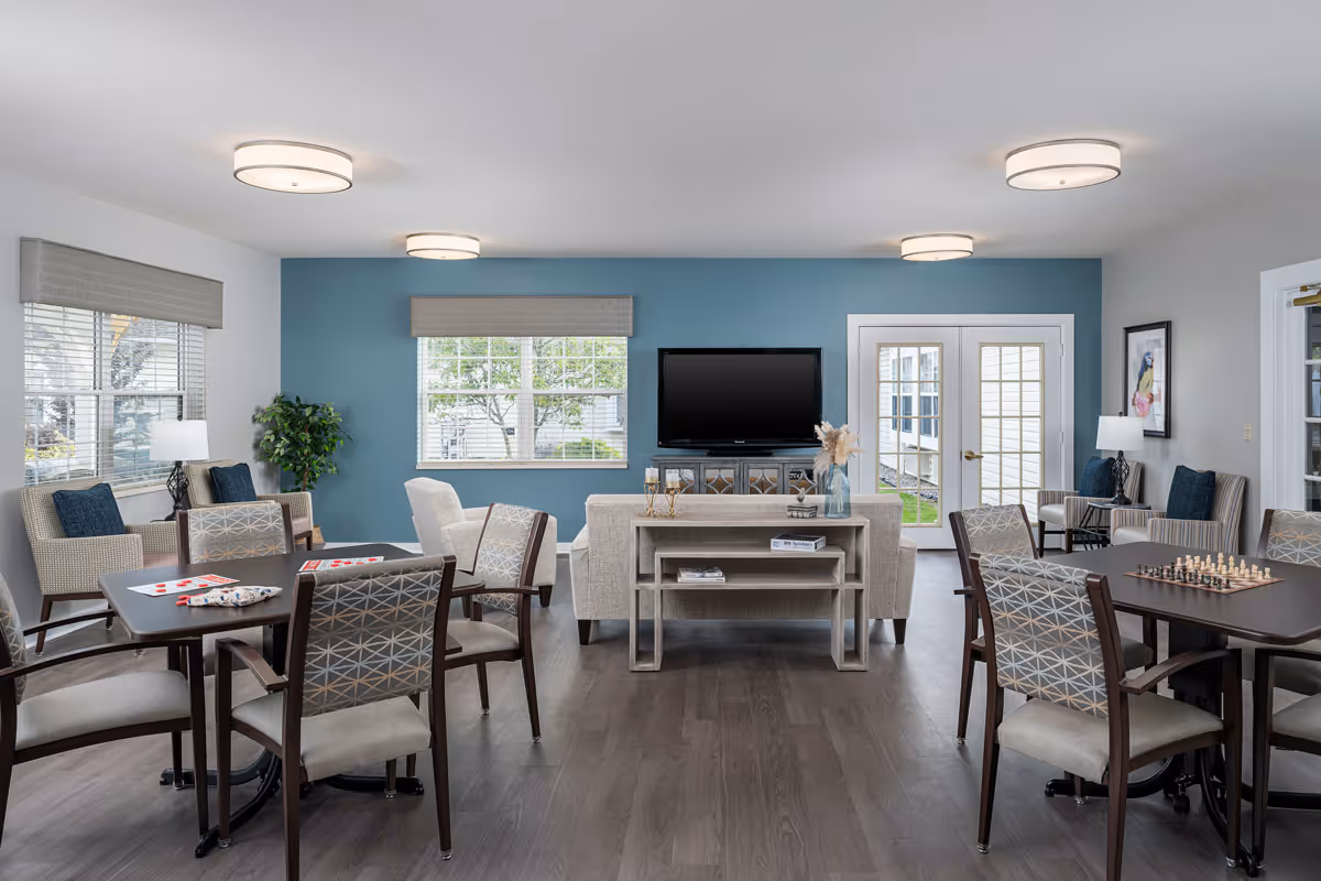 A bright and spacious common living area with multiple seating arrangements including armchairs and cushioned chairs around tables. The room features a blue accent wall with a flat-screen TV mounted above a console table. There are large windows with blinds, modern ceiling lights, and a chessboard on one of the tables, creating a welcoming and comfortable environment.