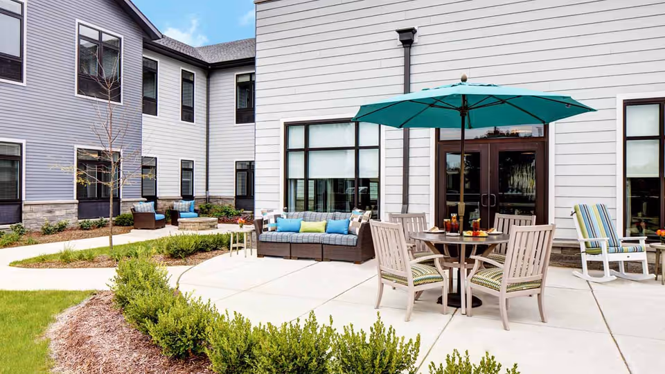 Outdoor patio area at StoryPoint Bolingbrook with a round table and four chairs under a large teal umbrella. There is a cushioned sofa with colorful pillows and additional seating including a striped rocking chair. The patio is surrounded by a garden with shrubs and small trees, and the building exterior features large windows and light-colored siding.
