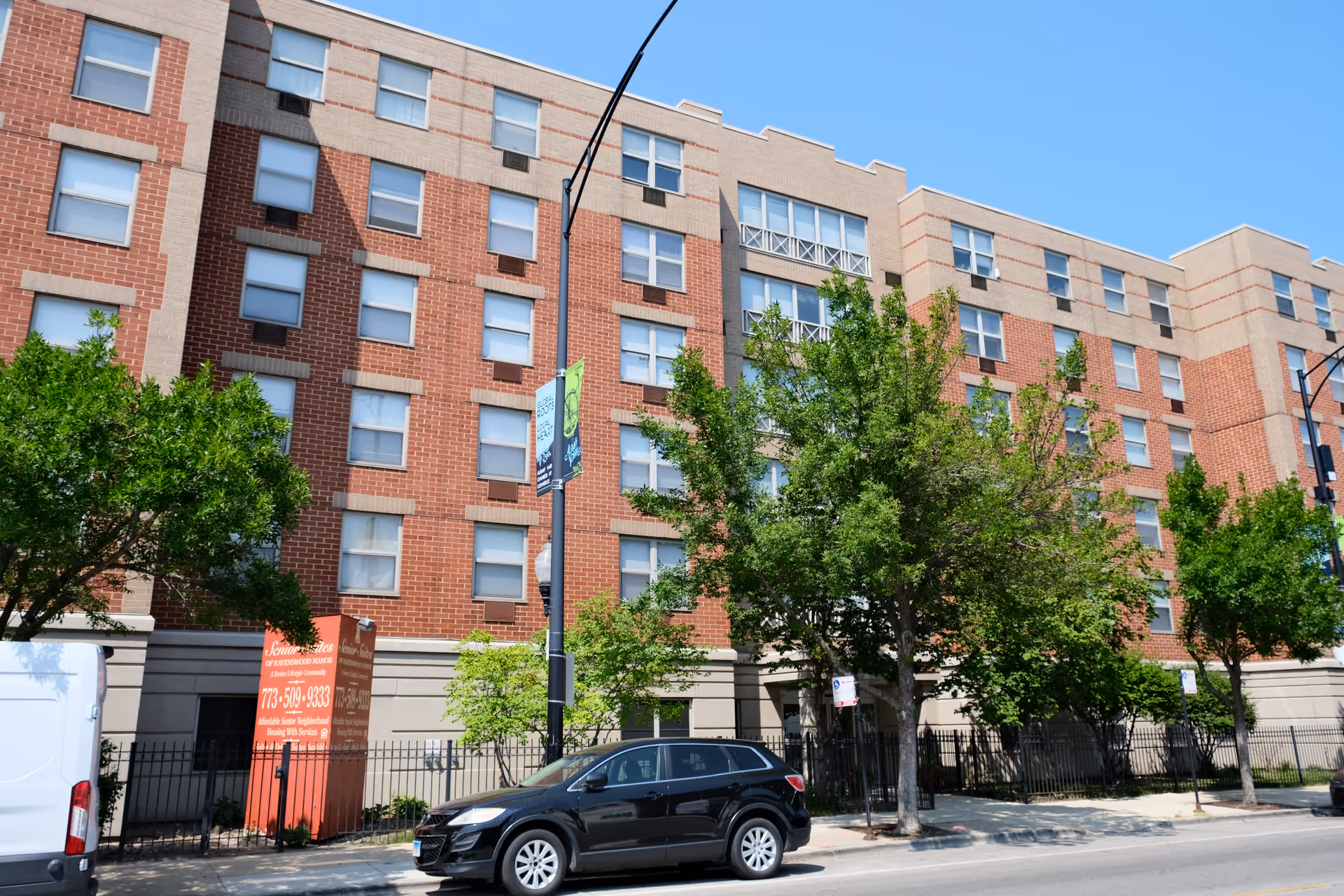 Front exterior of a multi-story red-brick senior living building with trees and parked cars along the street.