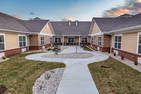 Outdoor courtyard area of a senior living facility with a concrete walkway, small landscaped garden beds with rocks and plants, surrounded by single-story buildings with beige siding, brick accents, and multiple windows under a partly cloudy sky at sunset.