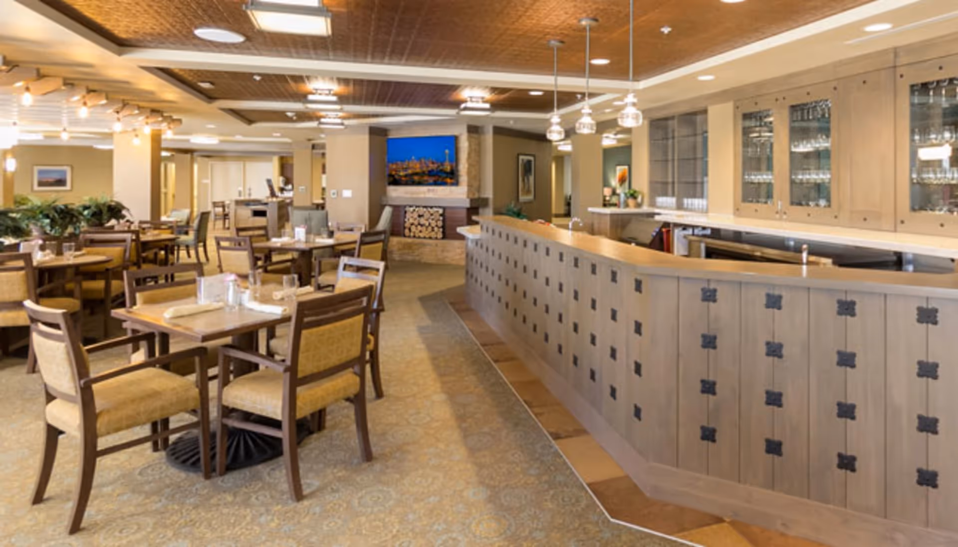 A spacious dining room with multiple wooden tables and chairs arranged neatly. The room features a long wooden counter with glass cabinets above it, pendant lights hanging from the ceiling, and a fireplace with a mounted TV above it in the background. The ceiling has decorative panels and the floor is carpeted with a patterned design.