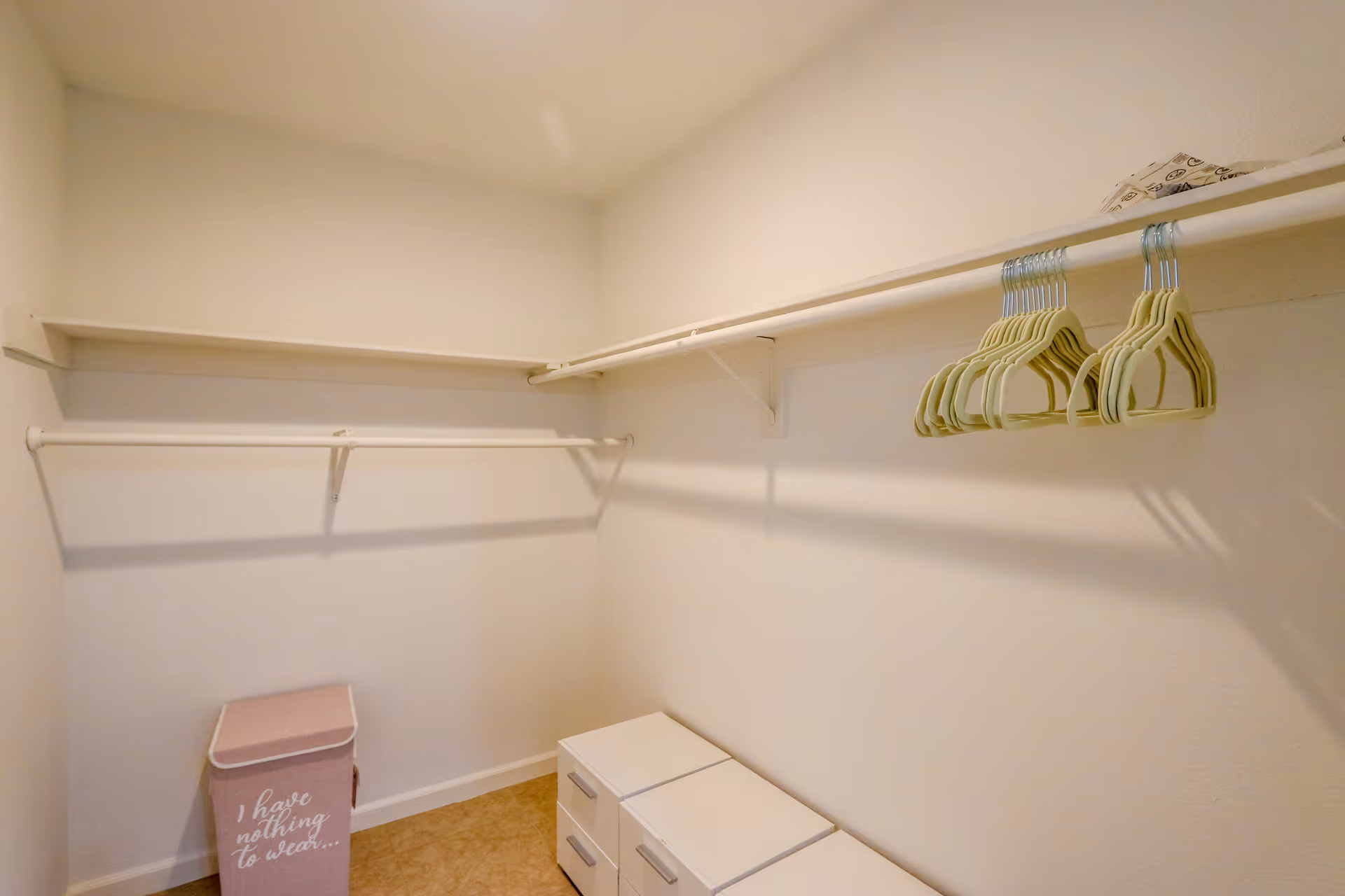 Empty walk-in closet with white walls, a row of beige hangers on a hanging rod, a pink laundry hamper with the text 'I have nothing to wear...', and three white storage drawers on the floor.
