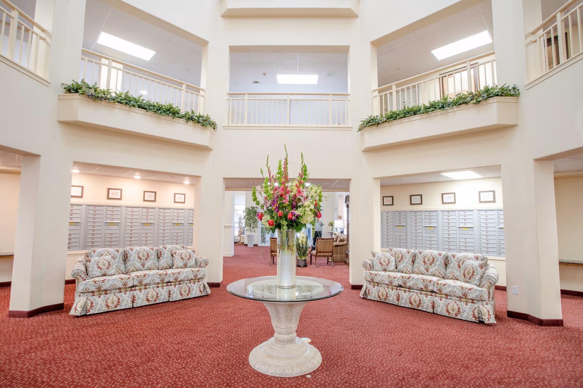 Atrium-style lobby with a central floral centerpiece on a round table, patterned sofas, and wall mailboxes.