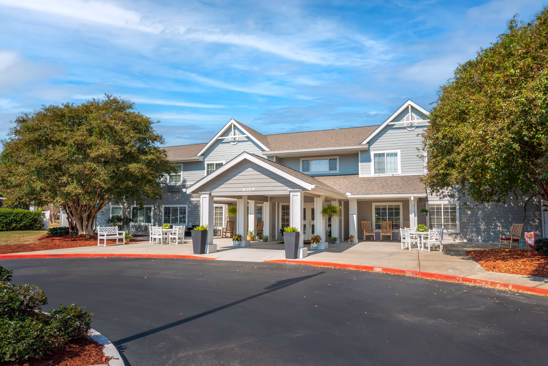 Exterior view of a two-story senior living facility building with a covered entrance, surrounded by trees and outdoor seating areas with white chairs and tables. The sky is partly cloudy with blue patches.