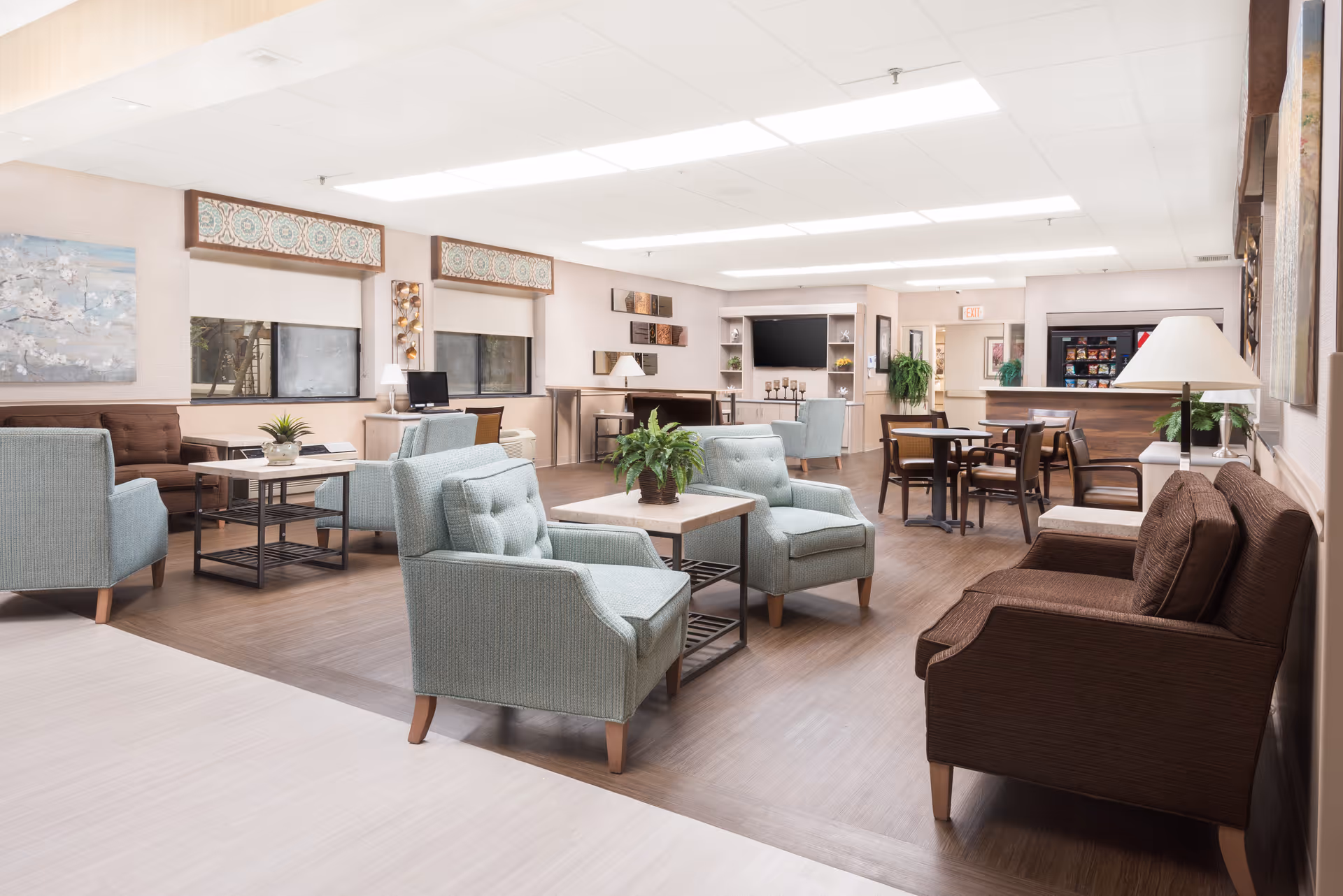 Well-lit communal lounge with armchairs, sofas, coffee tables, a TV and dining tables in a rehabilitation and healthcare center.