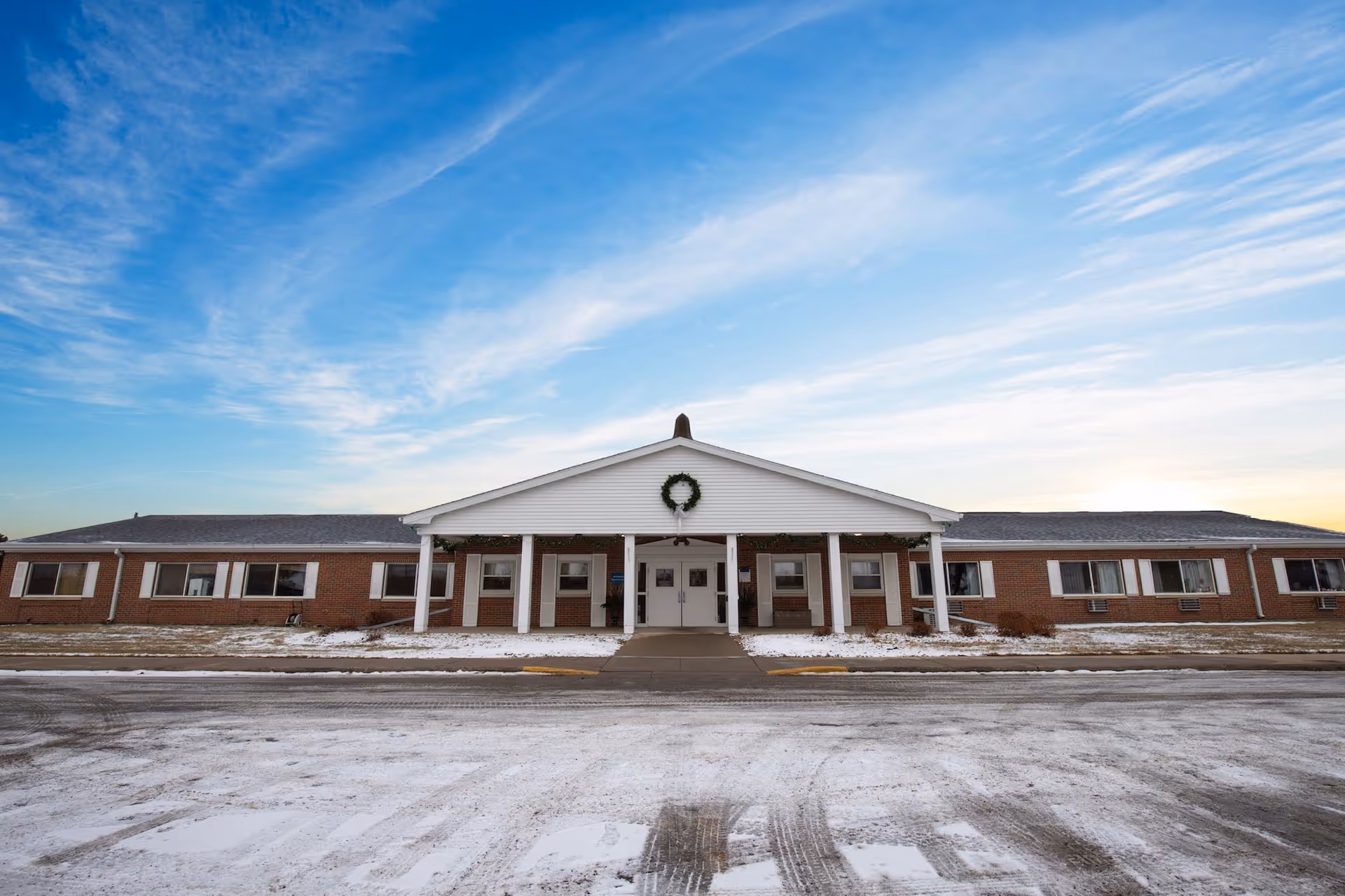 Front exterior view of Prairie Heights Healthcare building with a brick facade, white columns supporting a covered entrance, a wreath decoration above the entrance, and a partly snow-covered ground under a blue sky with scattered clouds.