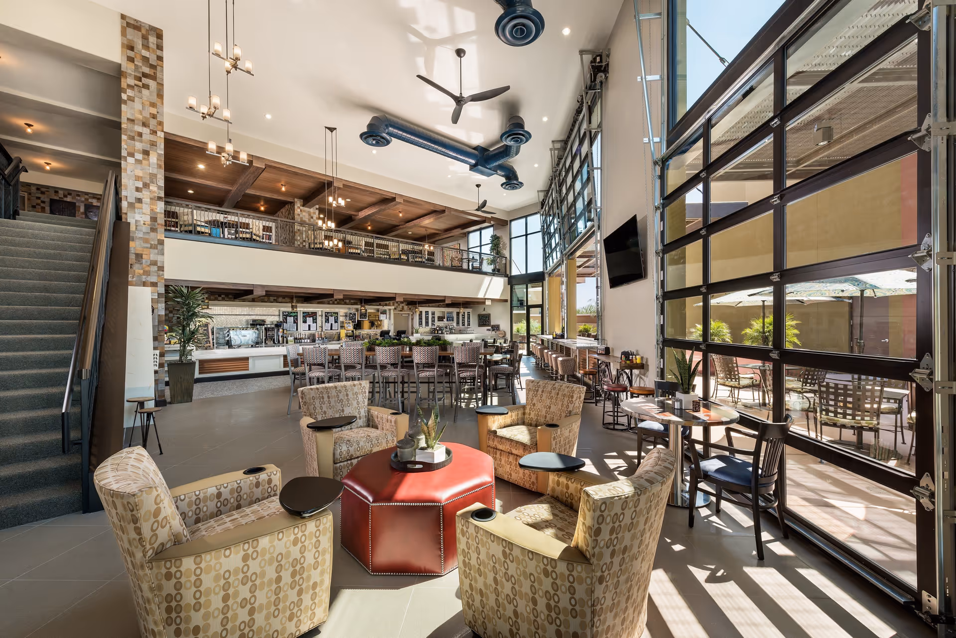 A spacious, modern common area with high ceilings and large glass garage-style doors letting in natural light. The room features a seating area with patterned armchairs around a red ottoman, multiple tables and chairs, a staircase leading to an upper level, ceiling fans, and a TV mounted on the wall. Outside, there is a patio with tables and umbrellas.