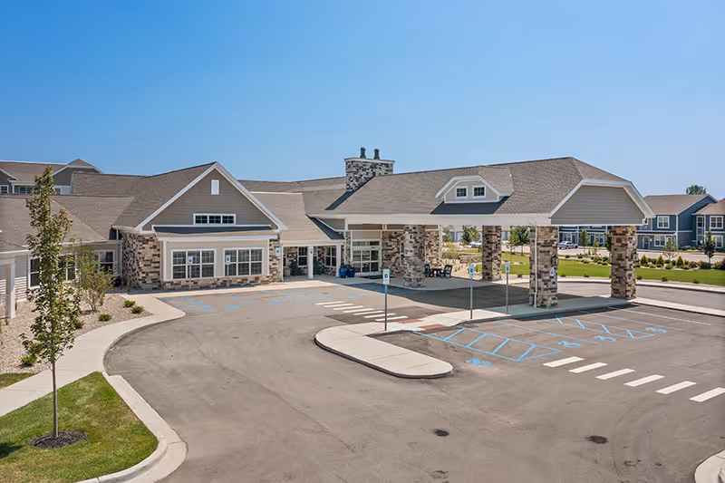 Exterior view of Quincy Place Senior Living facility showing a large covered entrance with stone pillars, a driveway with marked parking spaces including handicapped spots, and a clear blue sky.