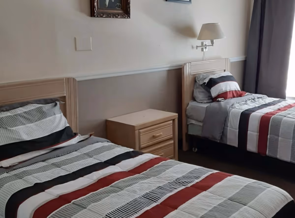 A bedroom with two single beds featuring striped bedding in shades of gray, black, white, and red. Between the beds is a wooden nightstand with two drawers. A wall-mounted lamp is above the nightstand, and a window with dark curtains is on the right side of the room.