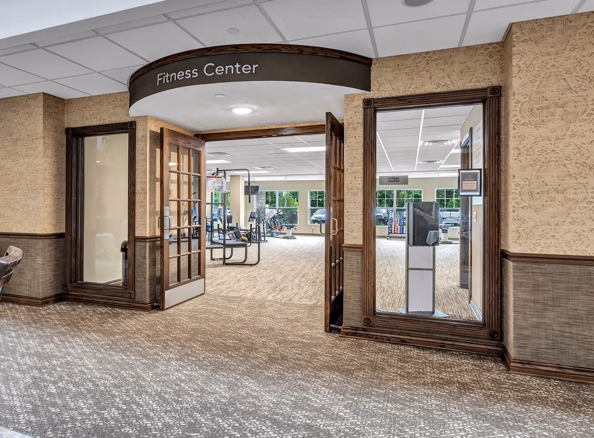 Entrance to a fitness center with double wooden doors open, showing exercise equipment inside. The area outside the fitness center has patterned carpet and walls with decorative wood trim and wallpaper. A large mirror is mounted on the right wall reflecting part of the fitness center.