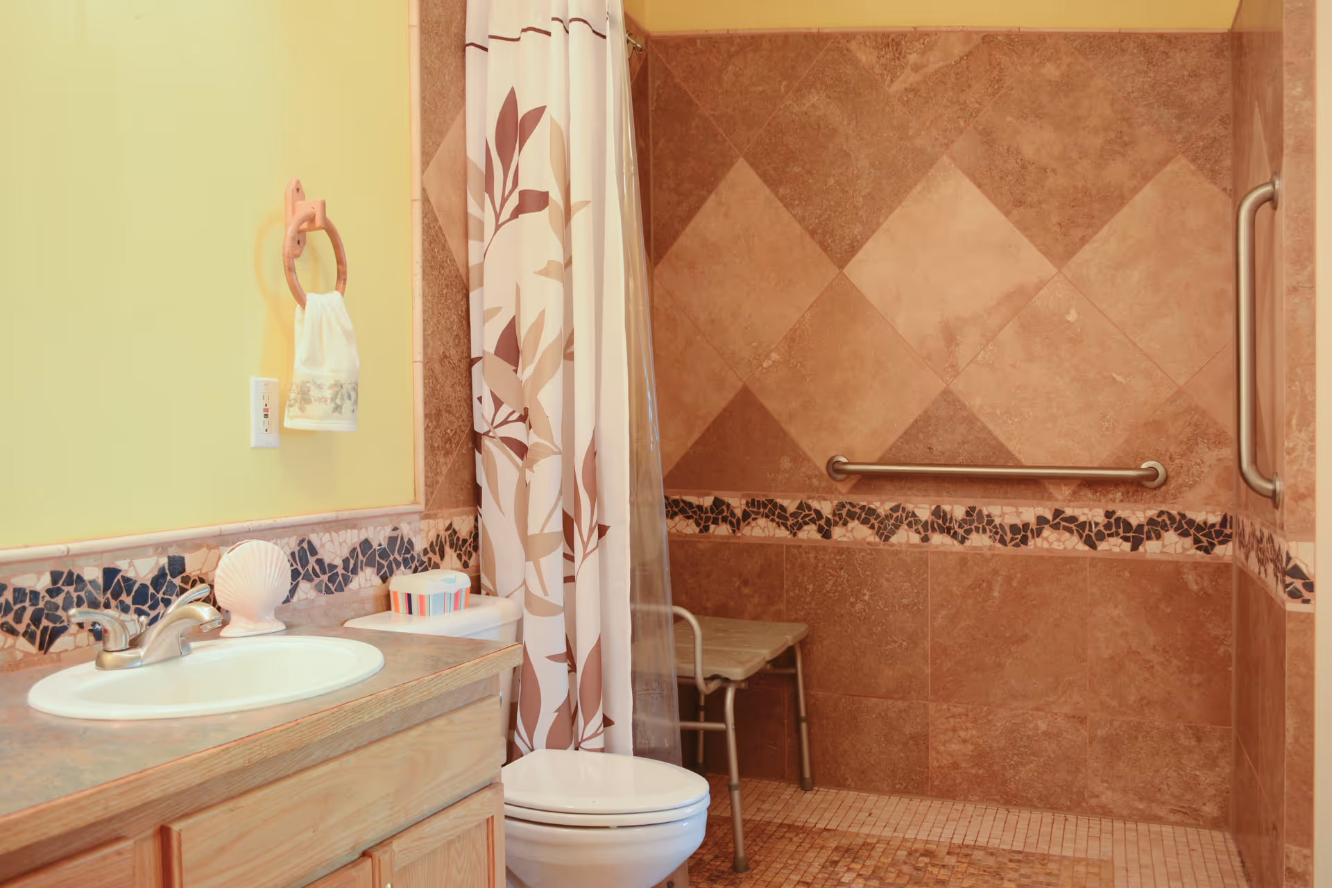 Bathroom with a sink and vanity, toilet, and a tiled walk-in shower with grab bars and a shower chair.